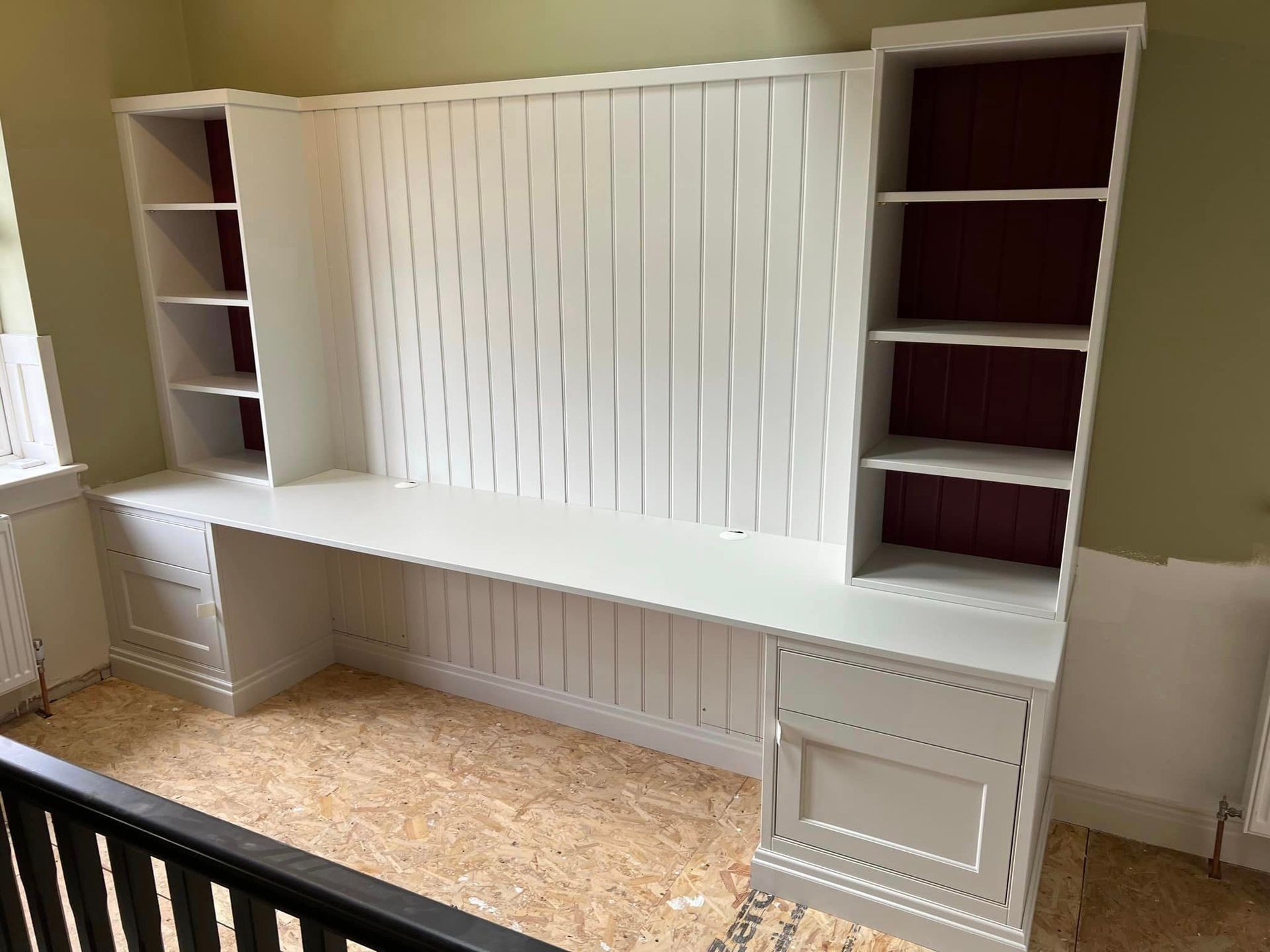 White built-in desk with shelves, set against a paneled wall in an unfinished room.