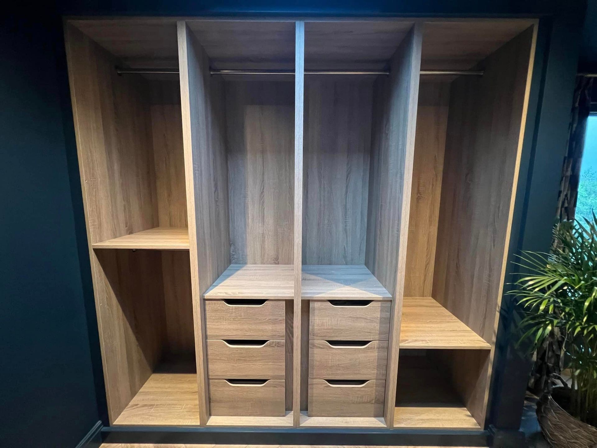 Empty wooden wardrobe with shelves and drawers, next to a potted plant.